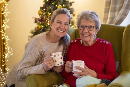 To kvinder, en ældre og en yngre, smiler, mens de holder krus foran et pyntet juletræ. Den ældre kvinde sidder i en grøn stol med garn i skødet, mens de deler ro og tryghed og samvær - ægte jul med demens-øjeblikke.