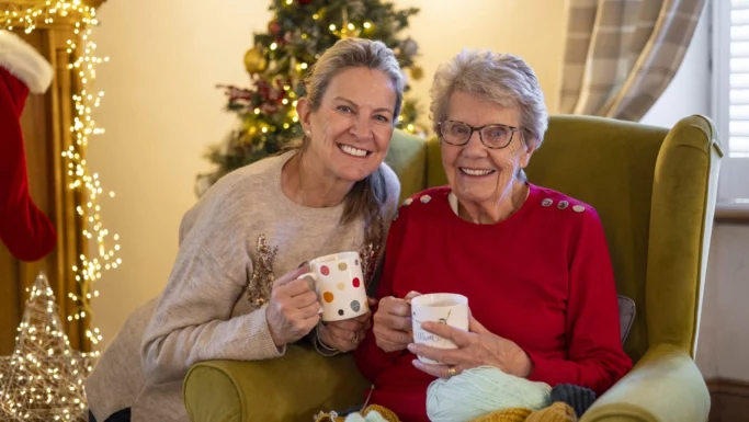 To kvinder, en ældre og en yngre, smiler, mens de holder krus foran et pyntet juletræ. Den ældre kvinde sidder i en grøn stol med garn i skødet, mens de deler ro og tryghed og samvær - ægte jul med demens-øjeblikke.