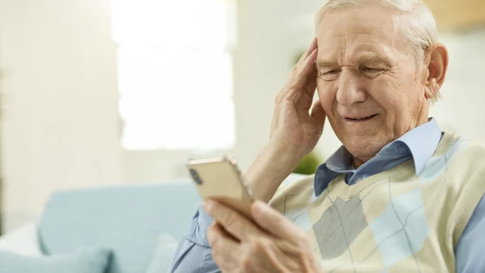 En ældre mand sidder i en sofa, holder en smartphone og rører ved sin tinding med et forvirret udtryk, muligvis kæmper han med digitalt valg i forbindelse med Ældrerådsvalg.