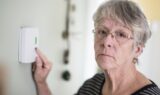 En ældre kvinde med gråt hår og briller trykker på en knap på en vægmonteret termostat og ser alvorligt på kameraet - måske overvejer hun stigende priser fra elselskaber eller elafgiftens indvirkning på hendes energiregninger.