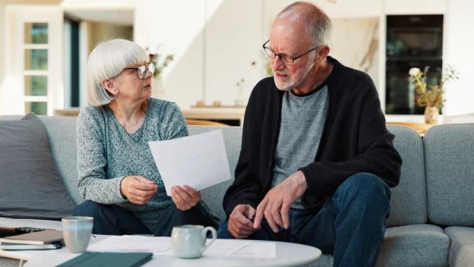 Et ældre par sidder i en sofa og gennemgår dokumenter sammen. Begge bærer briller og ser fokuserede ud. Papirer, en tablet og kaffekrus står på bordet foran dem i en veloplyst, moderne stue.