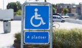 Et blåt og hvidt skilt viser tilgængelig parkering med et kørestolssymbol og teksten 4 pladser på dansk. Et handicapkort er påkrævet med en parkeringsplads og bygninger synlige i baggrunden.
