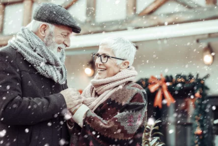Et ældre par i vintertøj smiler varmt til hinanden udendørs og holder hinanden i hånden, mens sneen falder - en hjertevarmende scene blandt juledekorationer og en bygning, der indfanger den festlige ånd, som mange danskere kender i løbet af sort jul.