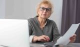 En ældre kvinde med briller sidder ved et skrivebord med en bærbar computer, holder et ark papir og bruger en lommeregner og ser fokuseret og eftertænksom ud, mens hun arbejder på sine IT-jobopgaver.