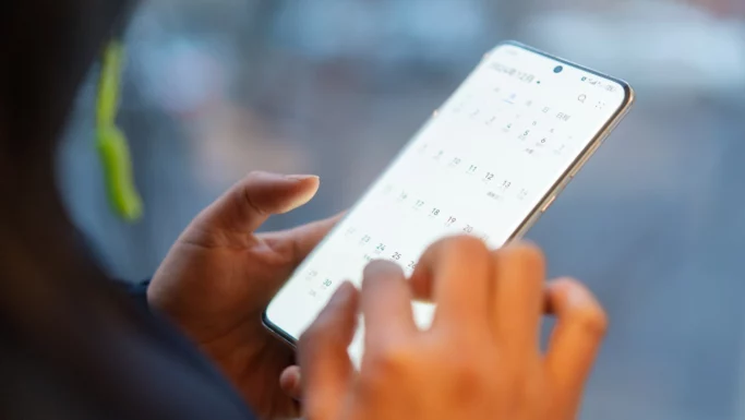 En person holder en smartphone og bruger fingeren til at scrolle gennem en digital kalender-app, hvor datoerne er tydeligt synlige på telefonens skærm, så man kan få stor hjælp til at organisere sine daglige planer.