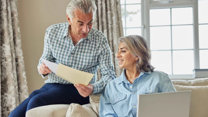 En ældre mand og kvinde sidder sammen på en sofa i en lys stue. Mannen holder og diskuterer papirer, mens kvinnen smilende ser på skinke. En bærbar PC er åben på fanget hendes - de bruger den som guide for at komme i gang med at investere.