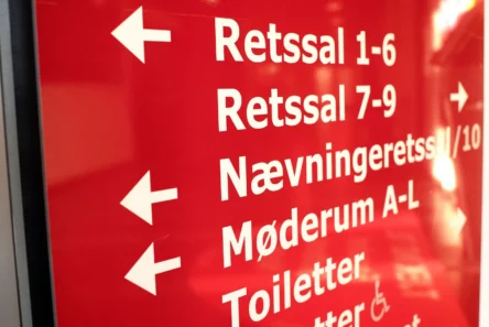 Et rødt skilt med hvid tekst og pile på dansk, der viser lægdommere, retspræsidenter, vej til retssalene 1-6, retssalene 7-9, nævningetinget, mødelokalerne A-L og toiletterne.