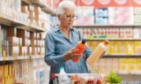 En ældre kvinde med kort hvidt hår og briller står i et supermarked og sammenligner to flasker - ligesom mange pensionister, der føler sig presset på økonomien, overvejer hun nøje sine valg. Hendes indkøbsvogn indeholder æbler, salat og drikkevarer på flaske.