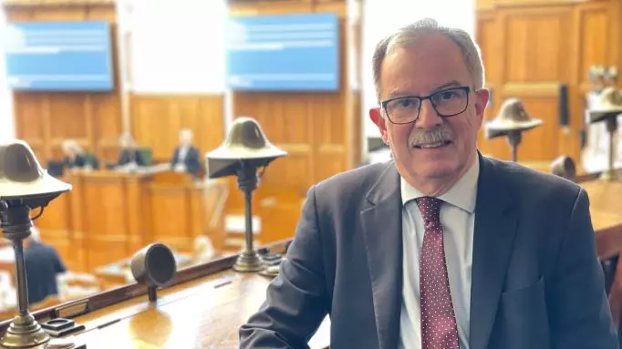 En mand i jakkesæt og prikket slips sidder i et træbeklædt rum - muligvis en retssal eller en lovgivende forsamling - hvor Peter Lautrup-Larsen engang diskuterede politiske blindgyder, med mennesker og store skærme slørede i baggrunden.