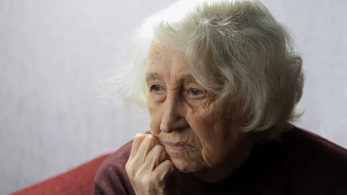 En ældre kvinde med hvidt hår hviler hagen på sin hånd og kigger eftertænksomt ud i det fjerne, mens hun reflekterer over sin partners død og er iført en rødbrun sweater. Baggrunden er svagt oplyst og ude af fokus.