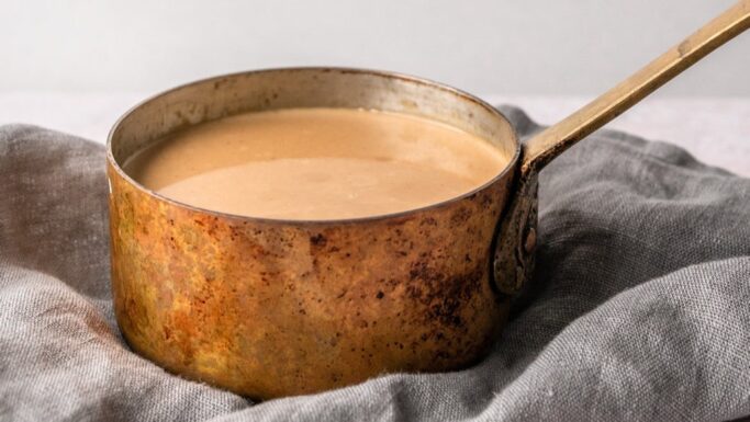 En rustik kobbergryde fyldt med lysebrun sovs står på et gråt klæde, perfekt til vegetariske juleretter, på en neutral baggrund.