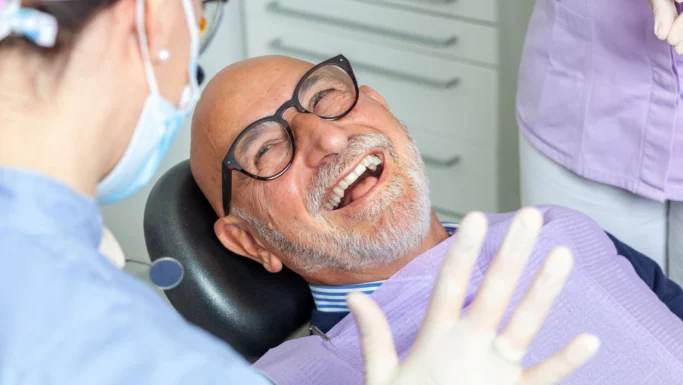 En ældre mand med briller og skæg smiler og griner, mens han sidder i tandlægestolen. En tandplejer iført håndsker og mundbind ses i forgrunden, hvor de sammen taler om tilskud til tandlægen.