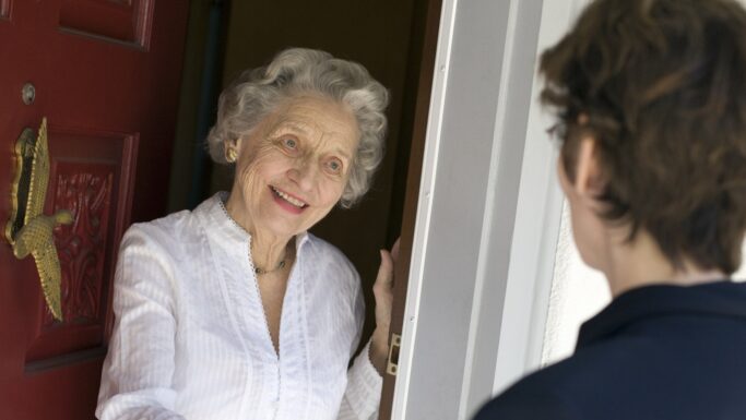En kvinde i 80'erne med gråt hår og hvid bluse smiler varmt, mens hun står ved sin røde hoveddør og sludrer med en teenager, hvis korte hår er synligt bagfra.