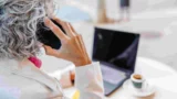 En ældre voksen med gråt, krøllet hår taler i en smartphone, mens han sidder ved et skrivebord med en åben laptop og muligvis diskuterer en fejl med sin tjenestemandspension, som kan kræve, at han betaler tilbage. Scenen ser ud til at være i et lyst indendørs rum.