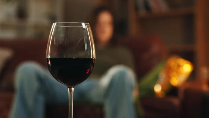 Et nærbillede af et glas rødvin i fokus, der fremhæver den afslappende atmosfære, men også minder os om risikoen forbundet med alkohol og potentielle sundhedsproblemer som hjerneblødning, med en person på en sofa i den svagt oplyste baggrund.