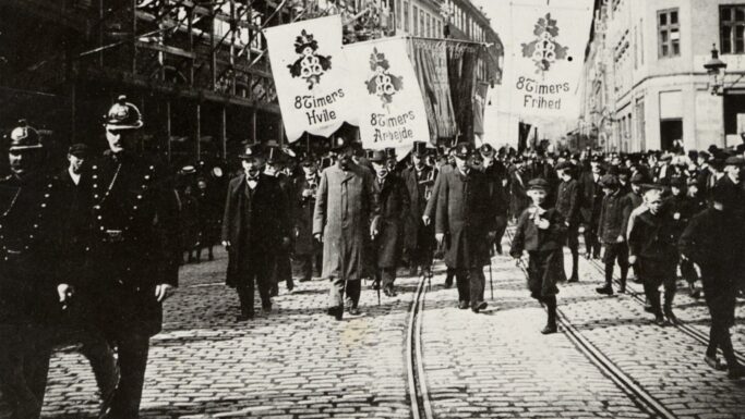 Sort-hvidt foto af en historisk gadeprotest, der afspejler arbejderhistorie, med mænd, børn og politi, der marcherer. På bannere på dansk står der 8 timers Hvile, 8 timers Arbejde og 8 timers Frihed (8 timers hvile, arbejde, frihed).