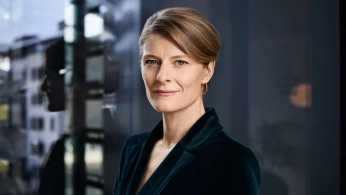 En kvinde med kort blondt hår iført mørk blazer og guldøreringe står indendørs og kigger selvsikkert ind i kameraet. Et bybillede spejler sig i vinduet bag hende og hentyder til hendes rolle som skatteminister, der diskuterer fødevarecheck.