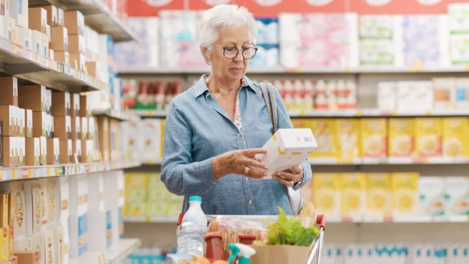 En ældre kvinde med kort, gråt hår og briller læser en etiket på en fødevarepakke og overvejer at gøre forskel på mødre, mens hun handler i en dagligvarebutik med en vogn fuld af varer.