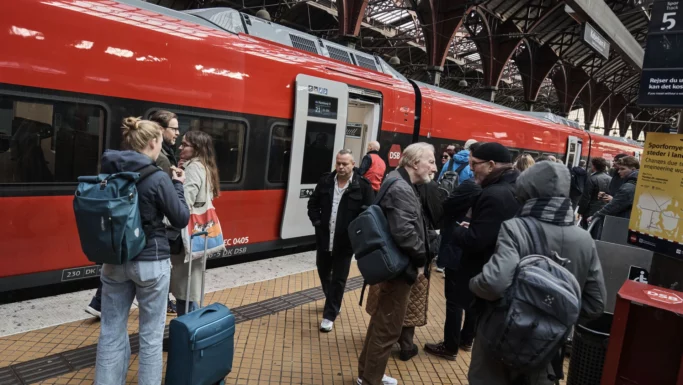 Folk med bagage står og snakker på en travl perron ved siden af et rødt DSB-tog, der forbereder sig på at stige på eller lige er ankommet, under et udsmykket stationstag - og nyder fordelene ved at bruge DSB-appen til deres rejse.