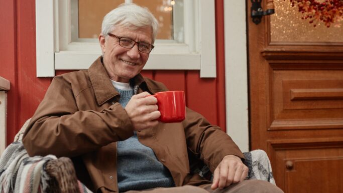 En ældre mand med gråt hår og briller sidder på en stol udenfor og smiler med et rødt krus i hånden. Han er iført en brun jakke over en blå sweater, og i baggrunden ses et rødt hus, som måske afventer nye ejendomsvurderinger.