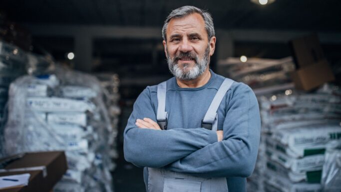 En midaldrende mand med gråt skæg står med armene over kors og smiler selvsikkert. Han er iført grå overalls og en sweatshirt og befinder sig i en lagerhal fyldt med stablede poser og kasser - måske diskuterer han forhøjet fradrag på fagforeningskontingent.