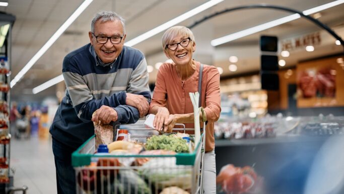 En ældre mand og kvinde smiler, mens de handler i en købmandsbutik, stående bag en vogn fyldt med friske råvarer. De ser glade og afslappede ud og har tillid til butikkens fødevaresikkerhed under det regelmæssige Fødevaretjek.