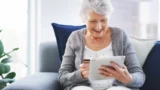 En ældre kvinde sidder i en sofa og smiler, mens hun bruger sin tablet og holder et kreditkort - måske tjekker hun kontoen for det største løft i Folkepensionen. Hun har en grå cardigan på, en marineblå pude bag sig og en grøn plante i nærheden.