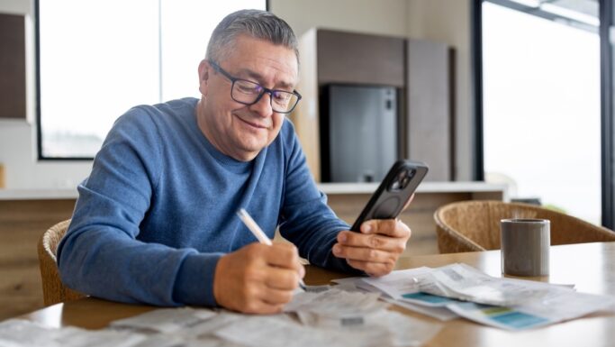 En midaldrende mand med briller og blå sweater sidder ved et bord og smiler, mens han kigger på sin smartphone. Han skriver på papir omgivet af kvitteringer og planlægger flere penge som pensionist med et krus i nærheden i et lyst, moderne køkken.