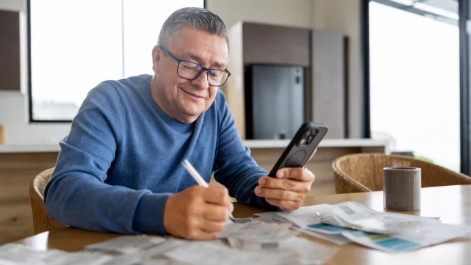 En midaldrende mand med briller og blå sweater sidder ved et bord og smiler, mens han kigger på sin smartphone. Han skriver på papir omgivet af kvitteringer og planlægger flere penge som pensionist med et krus i nærheden i et lyst, moderne køkken.