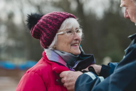 En ældre kvinde i rød frakke, tørklæde og strikket hue smiler varmt, mens en anden ældre person hjælper med at justere hendes frakke og forhindre varmetab fra hovedet på en kølig dag.