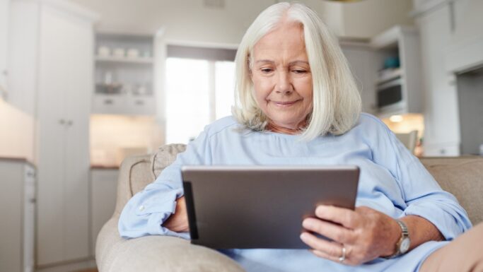 En ældre kvinde med langt gråt hår sidder i en sofa i en hyggelig stue og smiler, mens hun kigger på sin tablet og tjekker beskeder fra e-Boks.