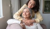 En ældre kvinde med Parkinsons sygdom sidder i en sofa og smiler, mens en yngre kvinde krammer hende blidt bagfra. De ser begge tilfredse ud i et hyggeligt, solbeskinnet rum.