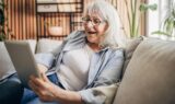 En ældre kvinde med gråt hår og briller sidder i en sofa og smiler, mens hun spiller skat på sin tablet. Hun ser glad og afslappet ud i en hyggelig, moderne stue med planter og hylder i baggrunden.