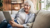 En ældre kvinde med gråt hår og briller sidder i en sofa og smiler, mens hun spiller skat på sin tablet. Hun ser glad og afslappet ud i en hyggelig, moderne stue med planter og hylder i baggrunden.