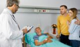 En læge fra sundhedspersonalet holder et clipboard ved en hospitalsseng og taler med en ældre mandlig patient. Hans pårørende - en yngre mand og kvinde - står ved siden af ham og trøster hinanden. En vase med blomster står på et bord i baggrunden.