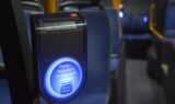 Nærbillede af en blå rejsekort-validator med dansk tekst på en bus. Bussen har blå sæder og gule stolper, mens den svagt oplyste baggrund skaber en rolig, afdæmpet atmosfære.