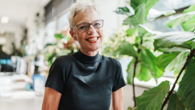 En smilende ældre kvinde, over 60 år, med kort gråt hår og briller står indendørs nær store grønne planter iført en sort kortærmet top. Klart naturligt lys falder ind gennem vinduer i baggrunden.