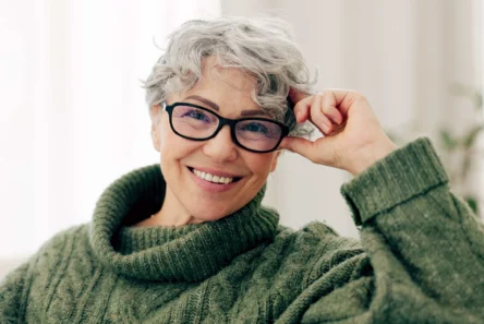 En ældre kvinde med kort, krøllet, gråt hår og briller smiler varmt iført en tyk, grøn sweater og sidder indendørs i et svagt oplyst rum.