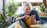 En ældre kvinde med gråt hår i hestehale sidder i en sofa, smiler og bruger sin smartphone til at tjekke MobilePay. Hun er iført en denimskjorte over en gul top, og der er stueplanter i baggrunden.