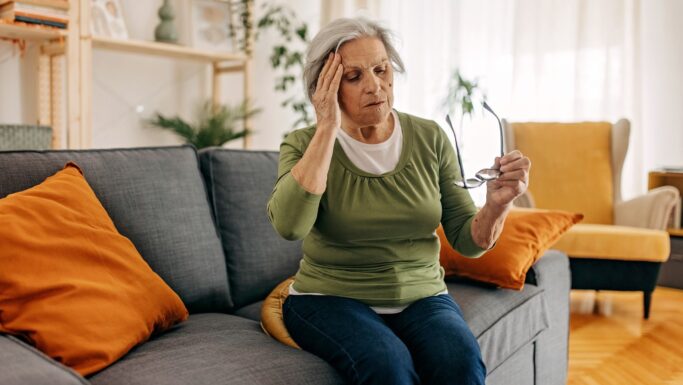 En ældre kvinde sidder i en sofa med sine briller i den ene hånd og rører ved sit hoved med den anden og ser ud til at føle sig utilpas og muligvis opleve svimmelhed. Det hyggelige rum har orange puder og indendørs planter.