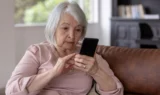 En ældre kvinde med gråt hår sidder i en brun sofa iført en lyserød top. Hun laver et telefonopkald på sin smartphone og bruger sine fingre til at interagere med skærmen. I baggrunden ses et lyst vindue.