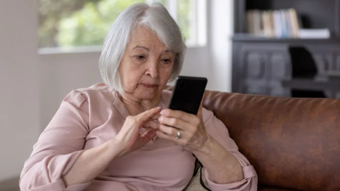 En ældre kvinde med gråt hår sidder i en brun sofa iført en lyserød top. Hun laver et telefonopkald på sin smartphone og bruger sine fingre til at interagere med skærmen. I baggrunden ses et lyst vindue.