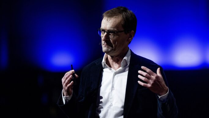 En mand med briller, en hvid skjorte og en mørk blazer taler på scenen om regulering med en headset-mikrofon. Han gestikulerer med hænderne, og baggrunden er dramatisk oplyst i blåt.
