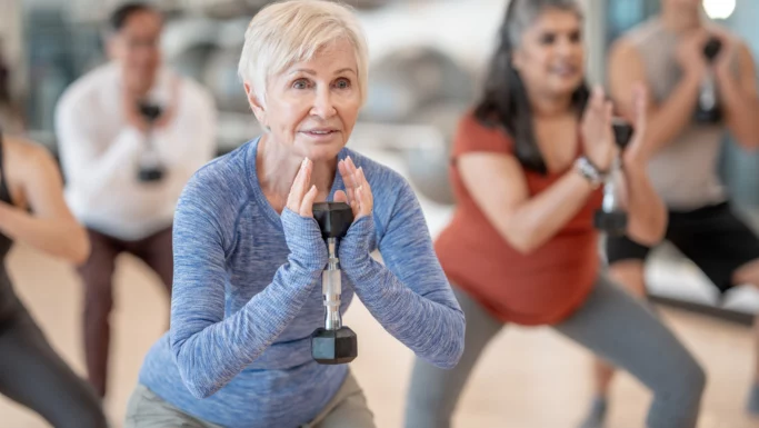 En gruppe mennesker i træningstøj udfører squats, mens de holder håndvægte i en fitnesstime. En ældre kvinde i en blå langærmet skjorte er i fokus og viser, hvordan få minutters bevægelse om dagen kan have stor effekt, ifølge ny forskning.