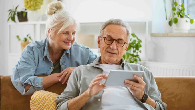 En ældre mand sidder i en sofa og bruger en tablet til at tjekke sin Årsopgørelse på skat, mens en ældre kvinde læner sig over skulderen på ham og smiler. De befinder sig i en lys stue med planter og moderne indretning i baggrunden.