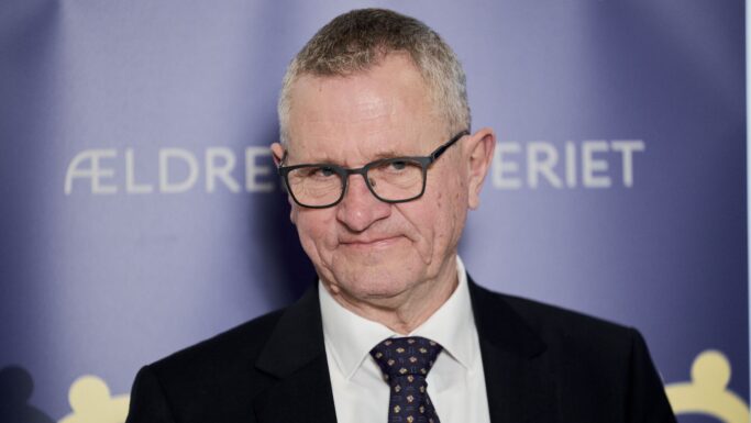 En ældre mand med briller, mørkt jakkesæt, hvid skjorte og mønstret slips står foran en blå baggrund med delvis synlig hvid tekst, hvor der står ÆLDRE og ERIET - muligvis under Henrik Frandsens valgkampen for Ældreministerposten.