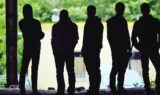 Fem personer står på rad og række, silhuetteret mod en lys udendørs baggrund med grønt og en gul bygning - muligvis relateret til svindel mod ældre eller anholdt i forbindelse med bandemiljøsager, deres træk er uigenkendelige.