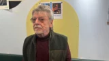 En ældre mand med gråt hår og briller står indendørs foran en opslagstavle med fotos og plakater. Visesangeren Benny Holst er iført en mørkegrøn vest over en rødbrun rullekrave. Den lyse baggrund supplerer udstillingen med musiktema.