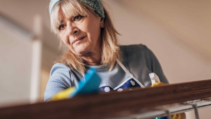 En pensionist med lyst hår iført blå bandana, handsker og forklæde rengør et trægelænder med en klud og en sprayflaske, mens hun koncentrerer sig om opgaven i et godt oplyst indendørs miljø.