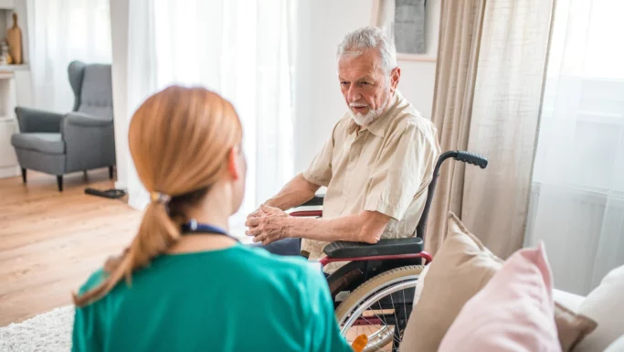 En ældre mand i kørestol sidder i en lys stue og taler med en sundhedsplejerske med rødligt hår om hjemmehjælp og dens indflydelse på hans hverdag.